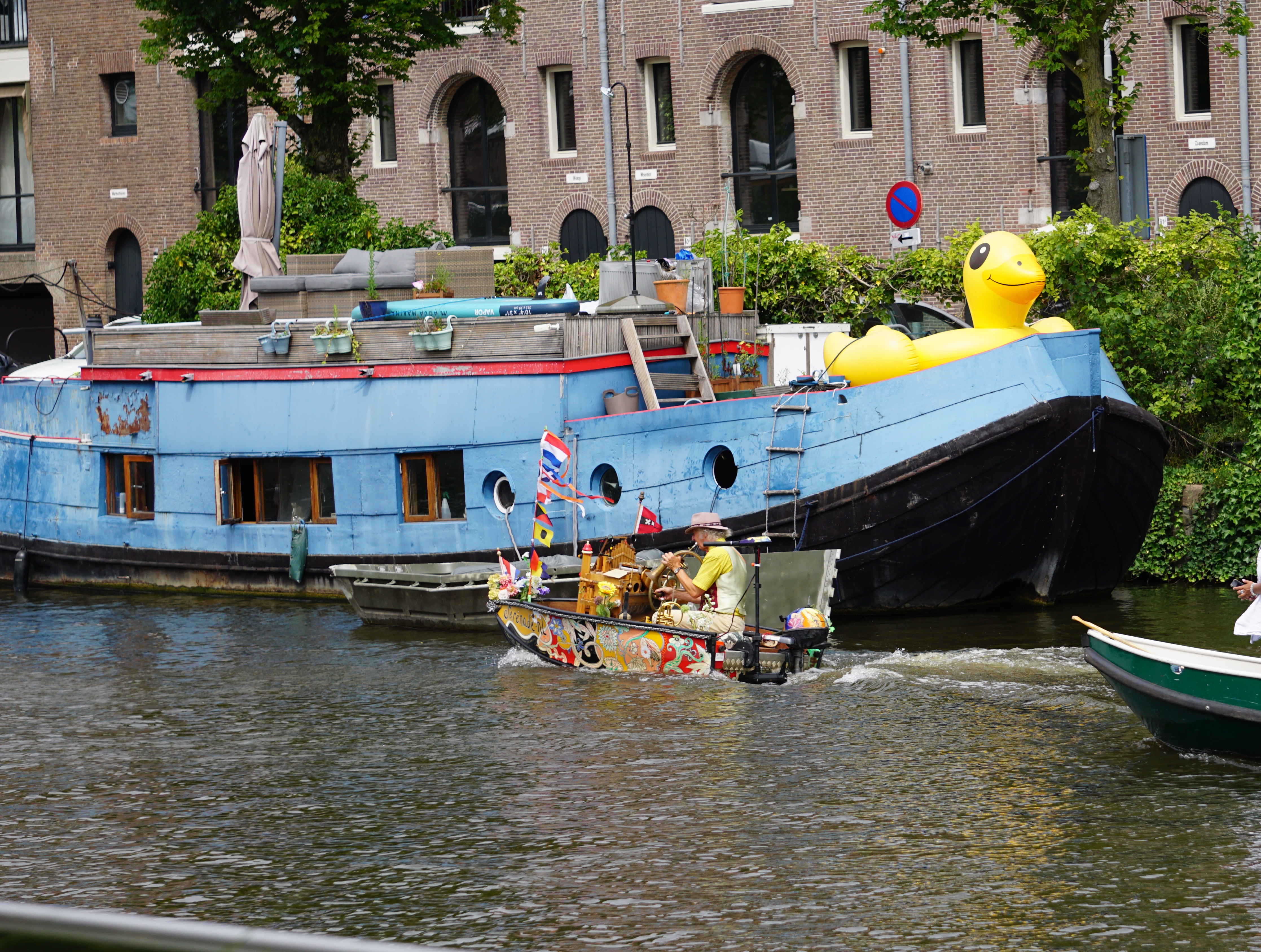 Hausboot im ganzen, davor ein kleines, bunt dekoriertes Boot mit Drehorgel, welches gerade daran vorbei fährt 