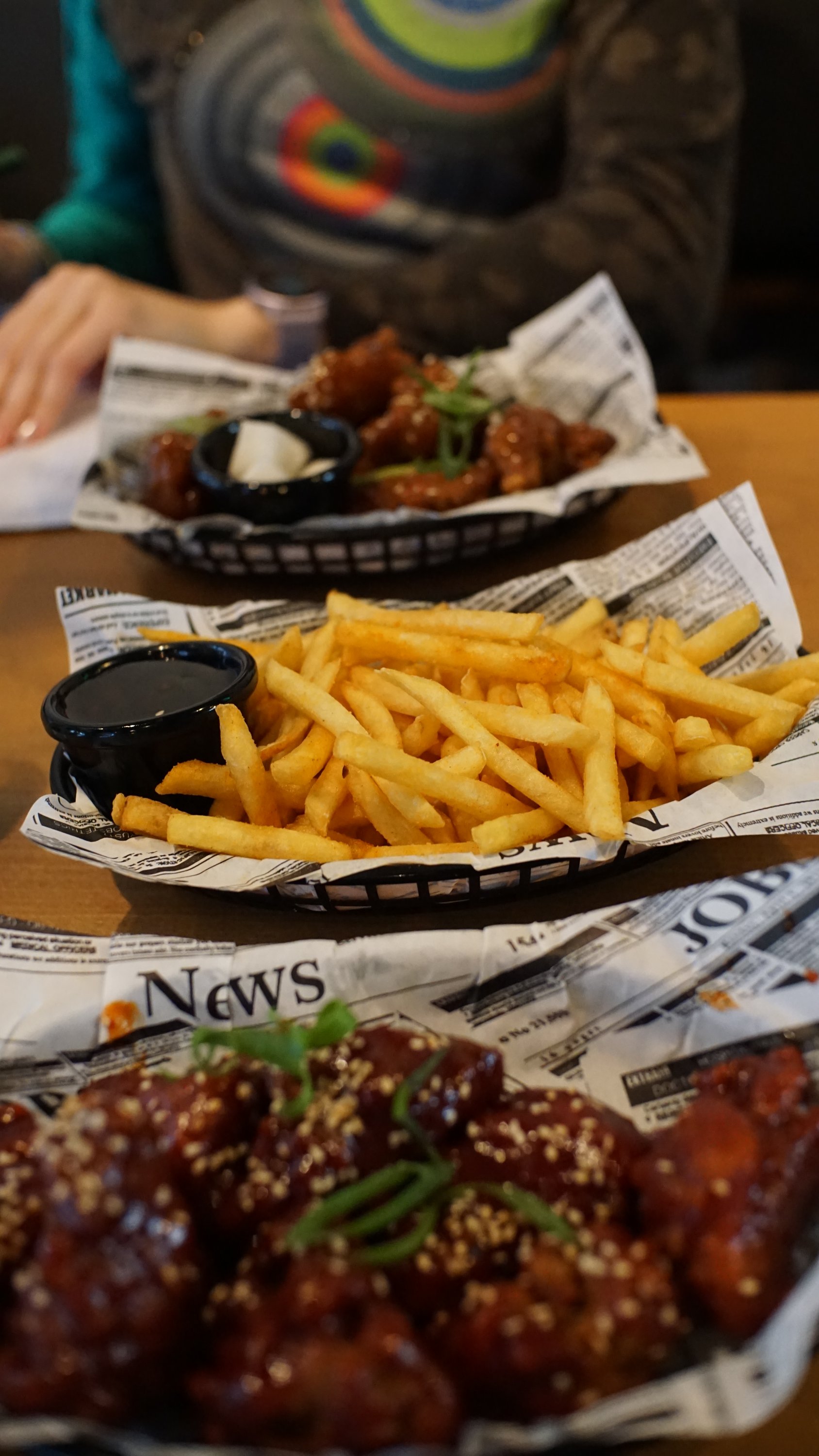 Tisch mit 3 Portionen Essen, jeweils fritiert und auf Papier in Körbchen 
Vorn im Bild sehr scharfes Hühnchen 
Mitte mit Pommes 
Dahinter weniger scharfes Hühnchen 

Fokus liegt auf den Pommes 