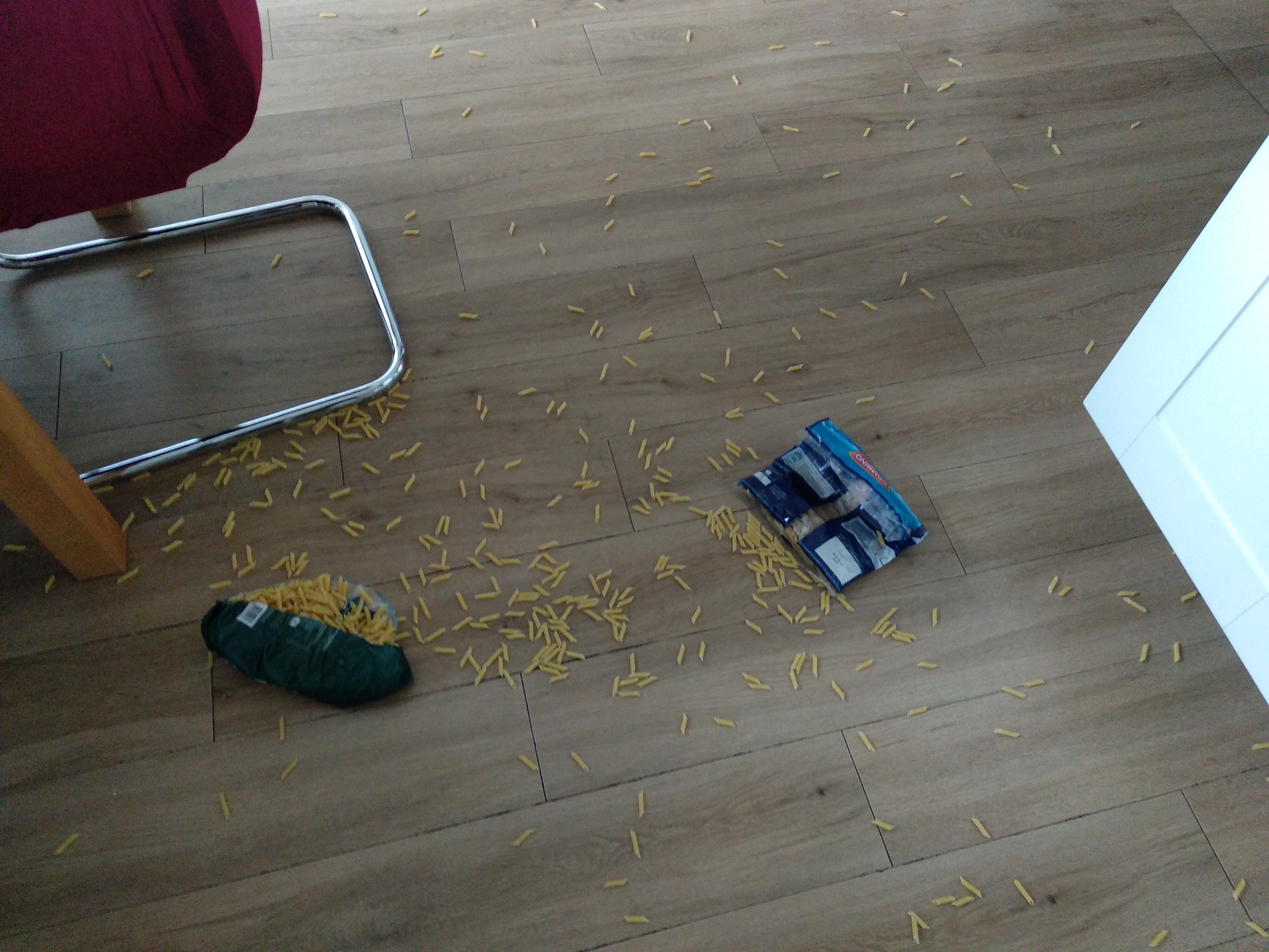 Küchenfußboden mit Laminat, vor einem offenen Schrank

Es liegen zwei offene/geplatzte Packungen Nudeln da

Überall sind Nudeln verstreut