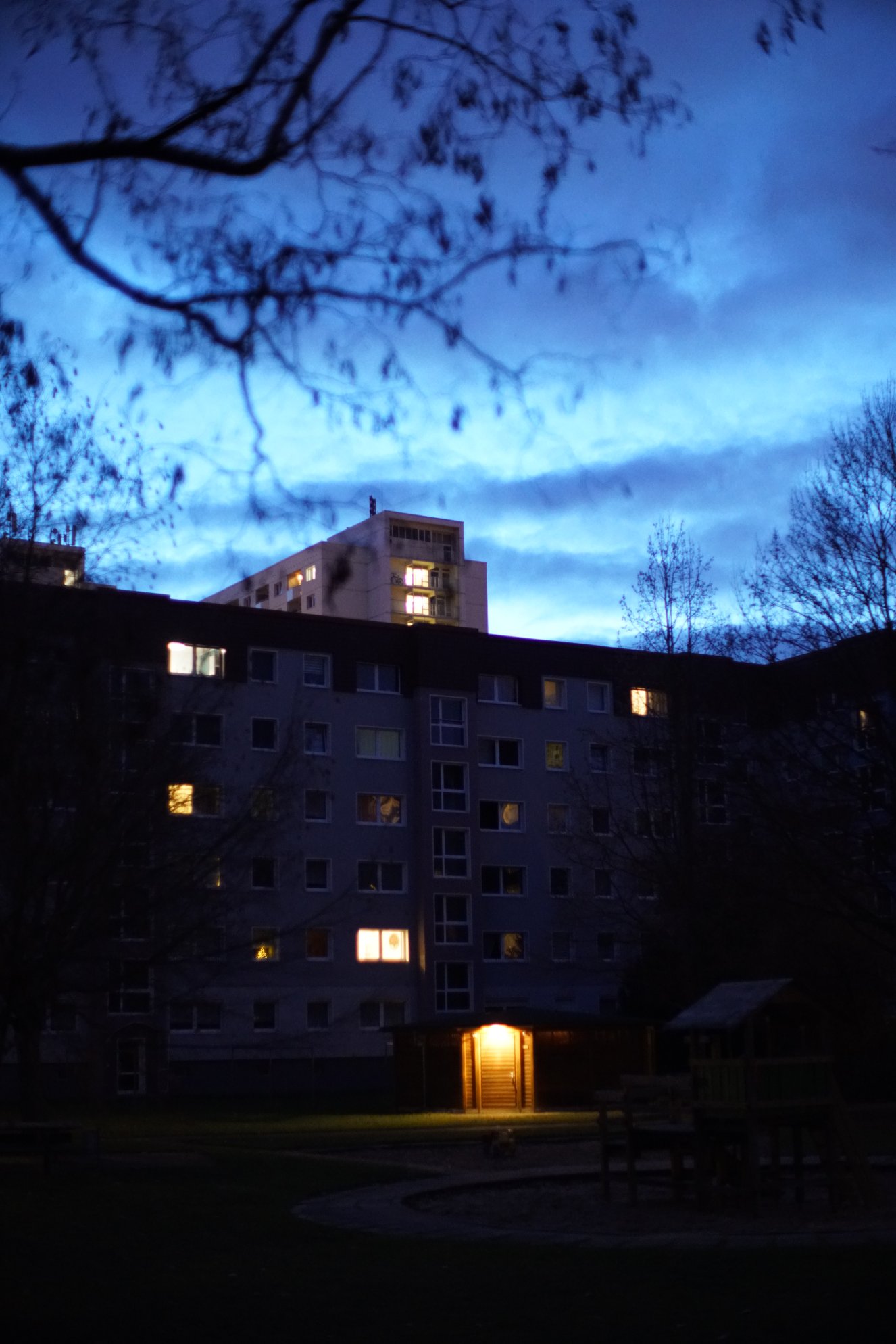Beton-Bau im Vordergrund, dahinter guckt ein höheres Gebäude ähnlicher Bauform darüber hinweg. Himmel dunkelblau, Zweige unscharf im oberen Bildrand.