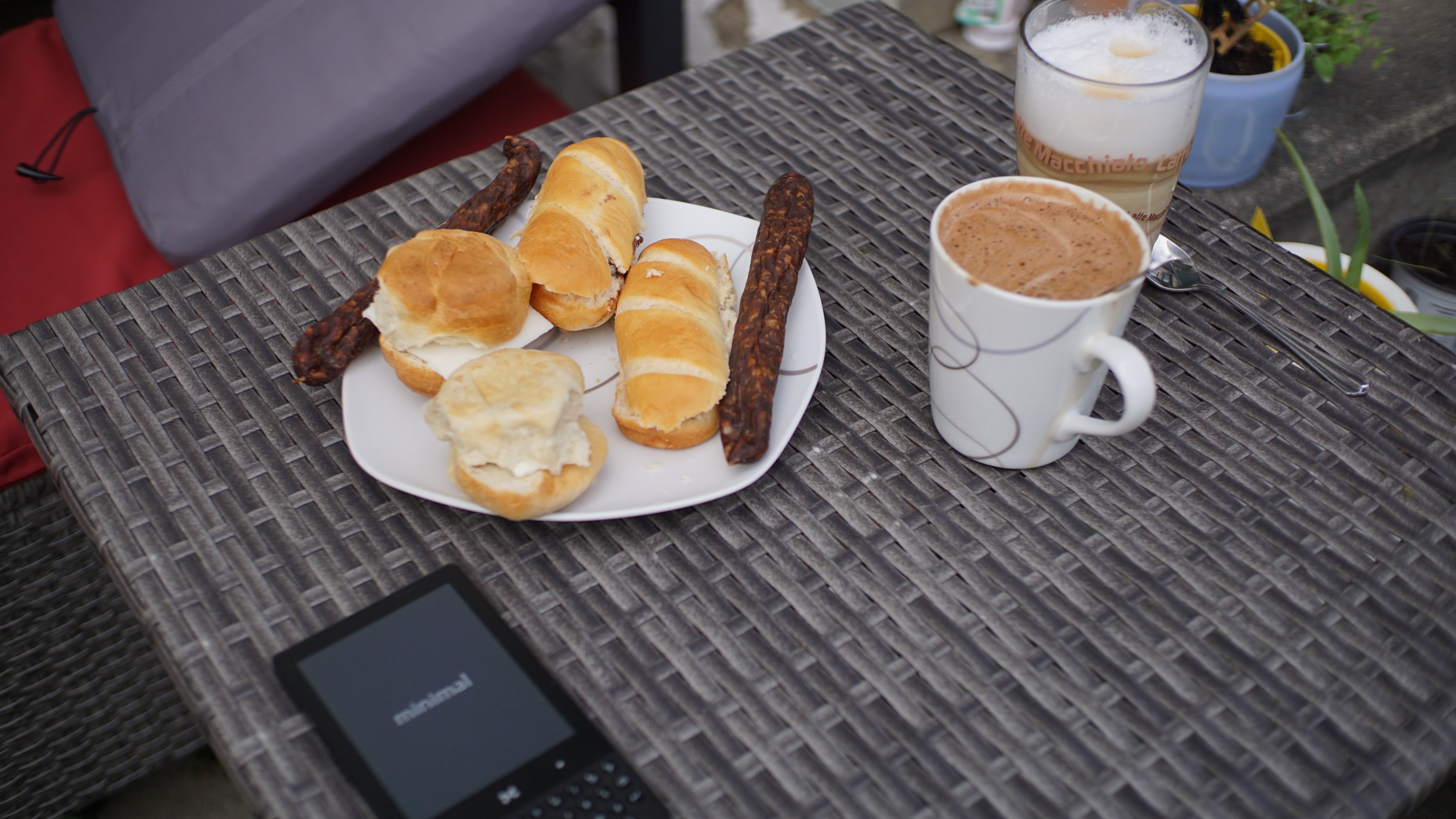 Milchbrötchen, kleinere Brötchen mit Käse und Lammknacker auf einem Teller 
Daneben zwei Tassen mit Kaffeegetränken und ein  "minimal"-Smartphone

Alles auf einem kleinen Gartentisch