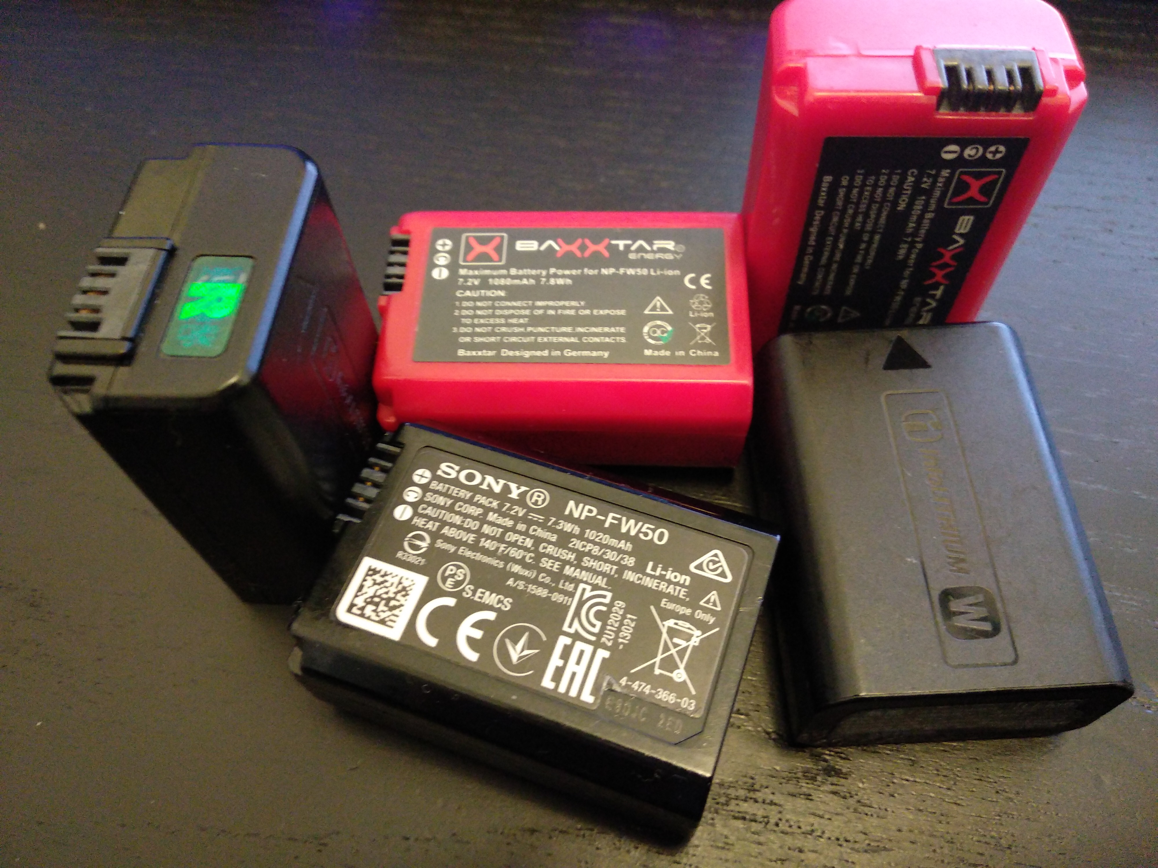 5 Akkus im gleichen Format auf einem Tisch.  3 davon 'original', mit Hologramm um das zu beweisen. 2 hingegen in rotem Gehäuse, von dritthersteller.