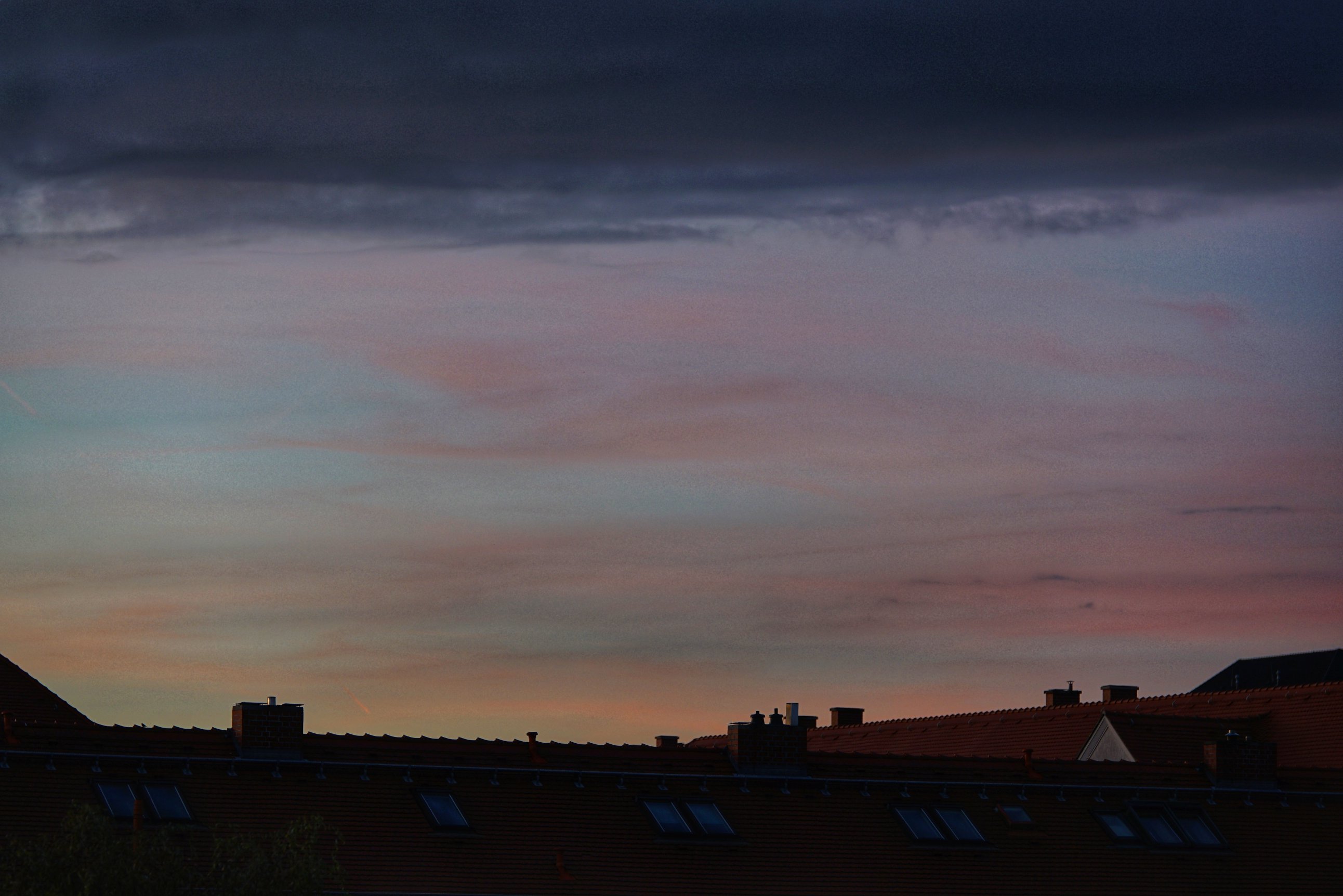 Wolkenbänder in unterschiedlichen Schattierungen, unten orange, dann rosa/rot, oben lila bis dunkelblau und schwarz.

Darunter Dächer 