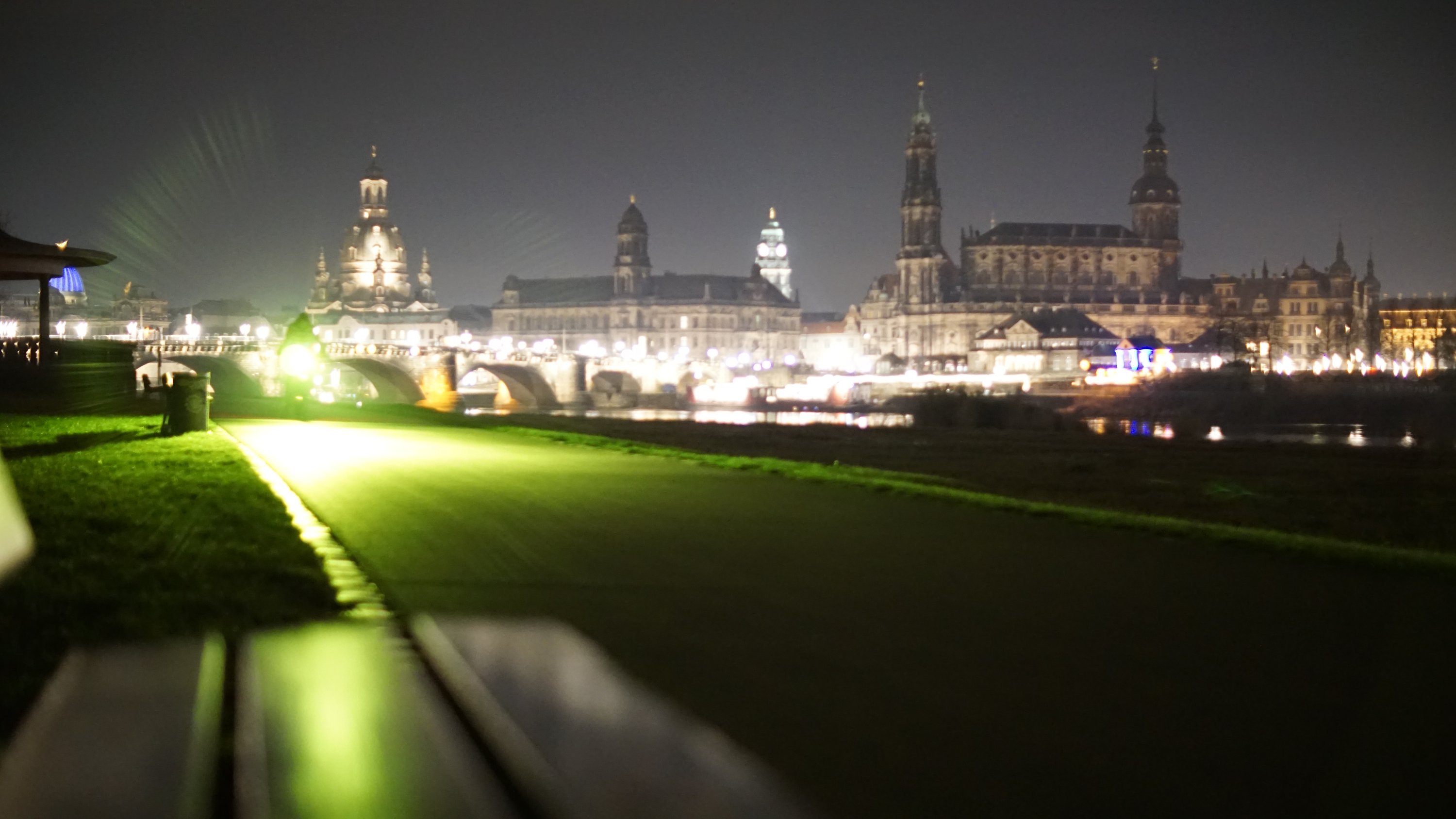 Blick über eine Bank, den Elberadweg entlang, zur Altstadt 
Radfahrer mit grell-grünem Licht kommt entgegen 
Altstadt im Hintergrund beleuchtet 