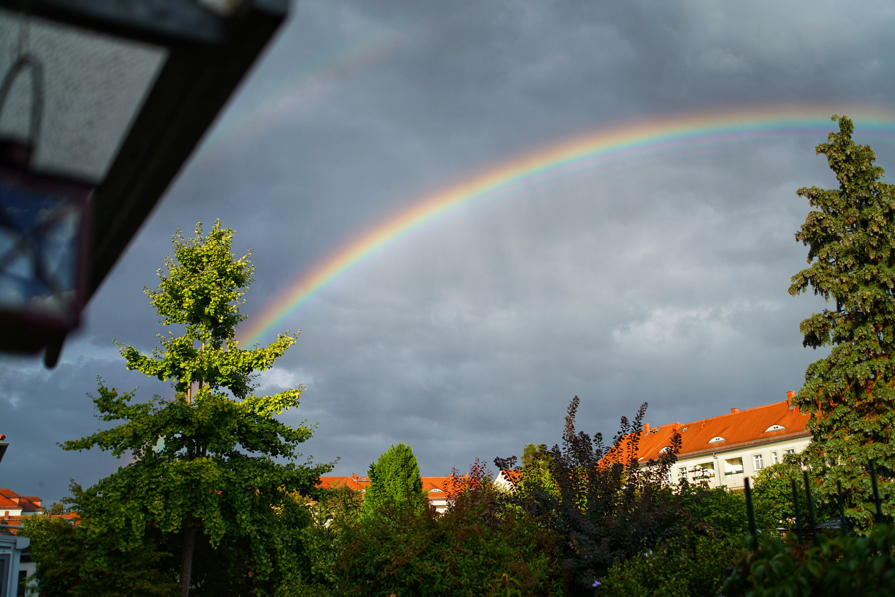 Regenbogen über Gärten 
Laterne am linken Bildrand 