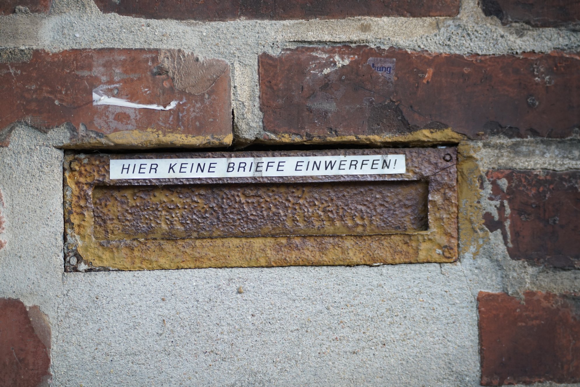 Alter, verrosteter Briefschlitz in einer Mauer, darüber Aufkleber "hier keine Briefe einwerfen!"