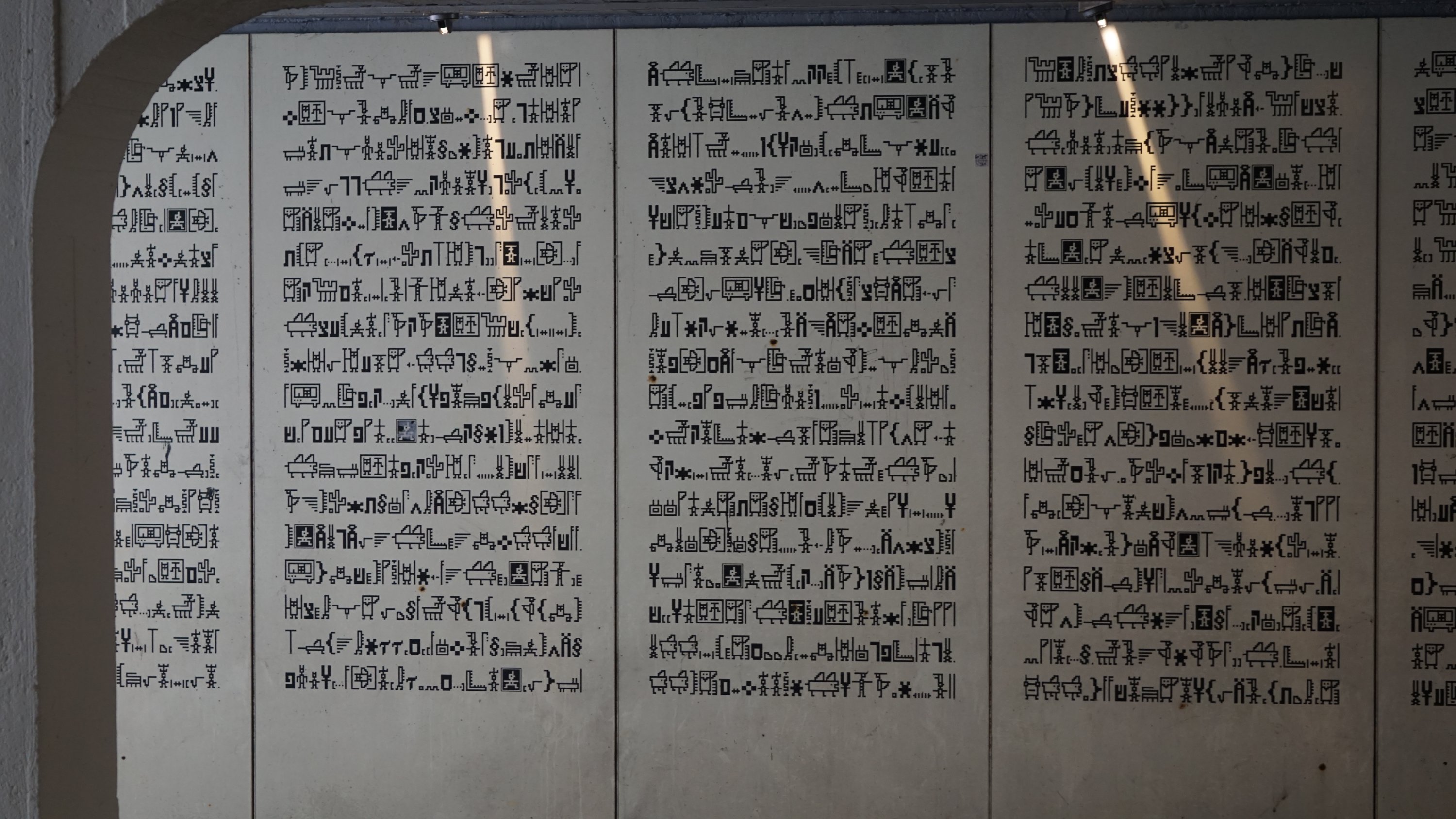 Wand voller Hieroglyphen, die einzelnen glyphen sind recht nah an Pixelart-eckig und stellen moderne Dinge wie Bahn, Auto, Badewanne oder Kakteen dar.

Jeweils ca. Zwei Dutzende Symbole breite Spalten, mindestens 5 solcher Spalten sind auf der Wand zu sehen. 