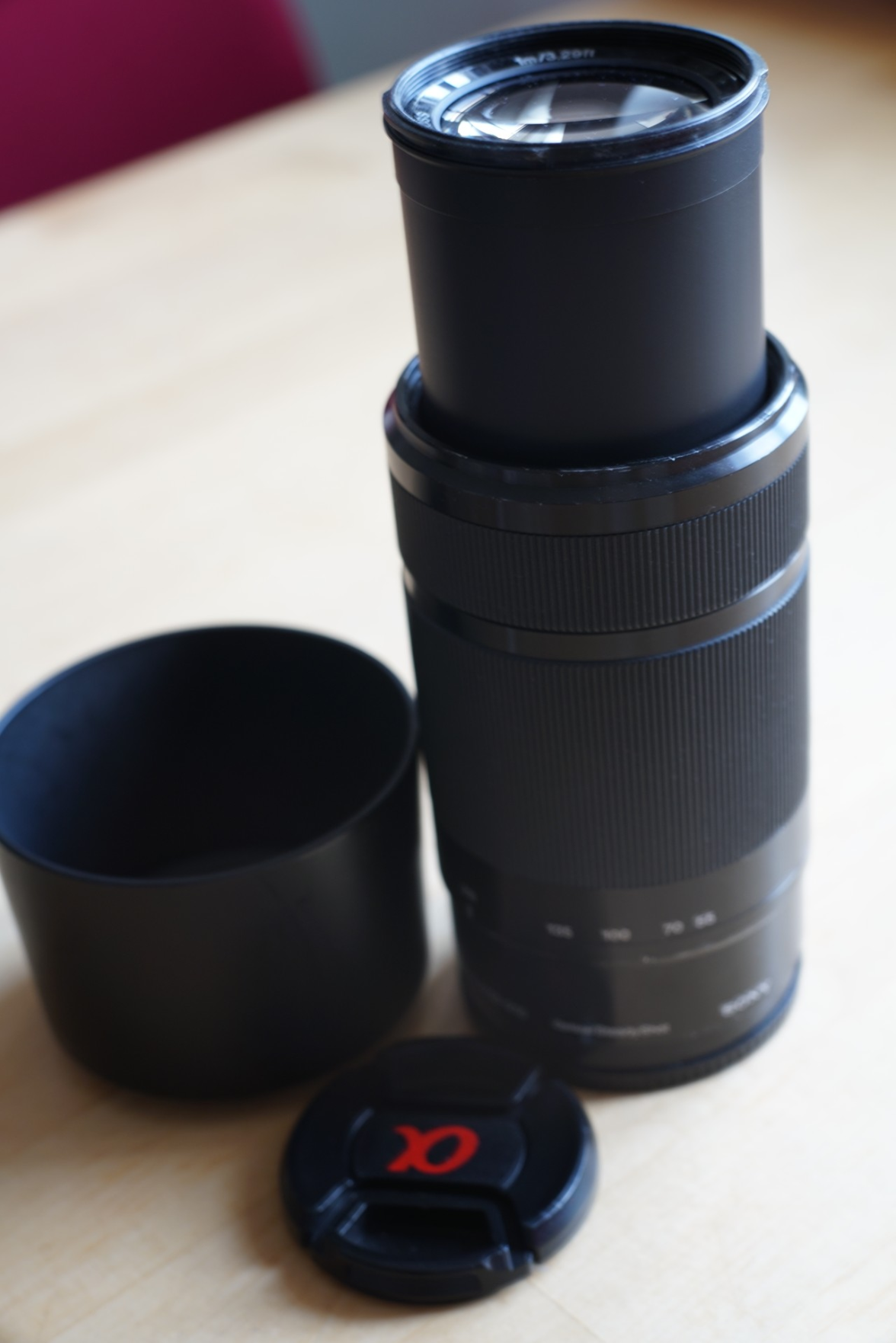 das ausgefahrene Objektiv, daneben die zugehörige Sonnenblende und ein Deckel.