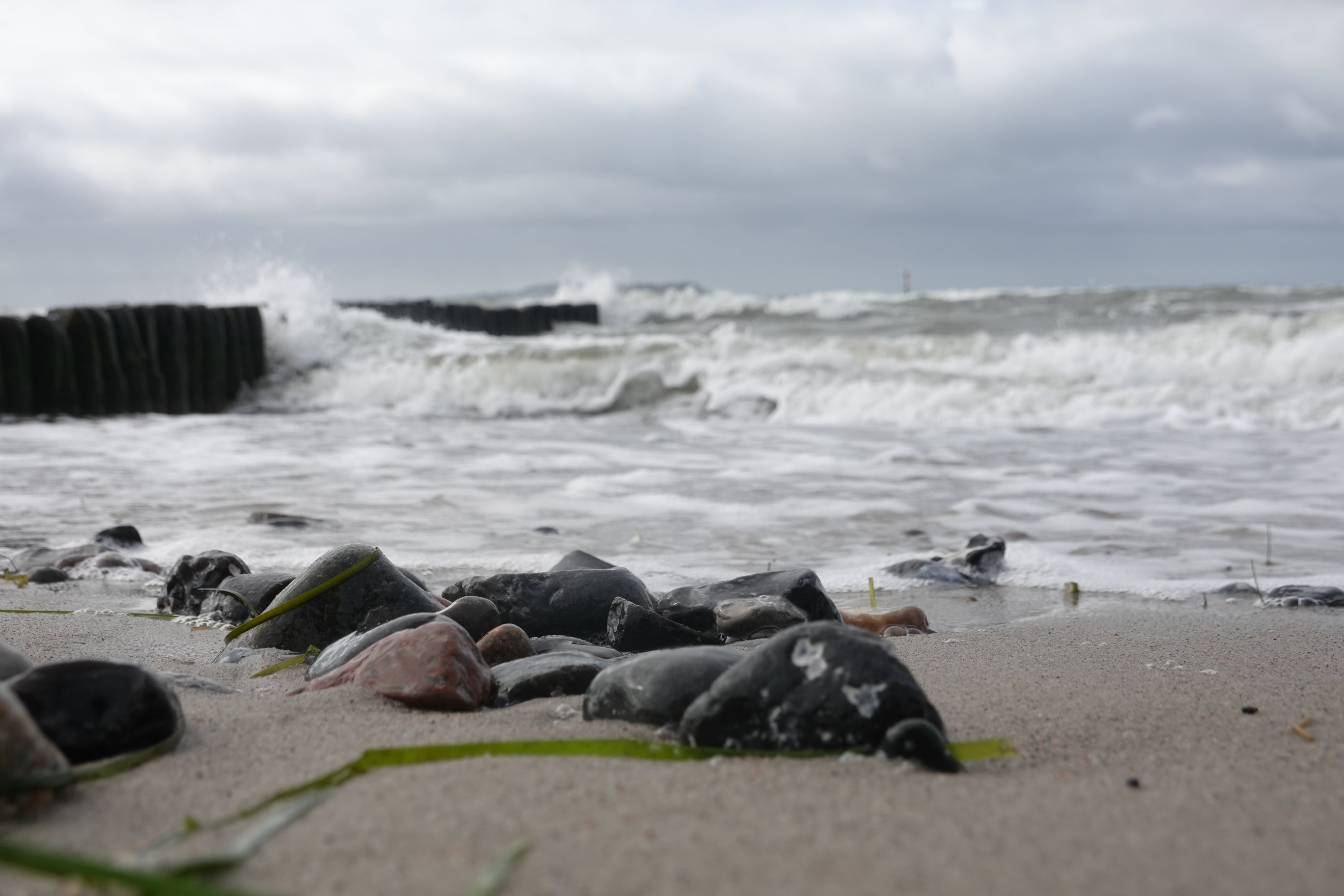 Mehr Steine zu sehen, im Hintergrund bricht sich eine Welle an den Holzbohlen 