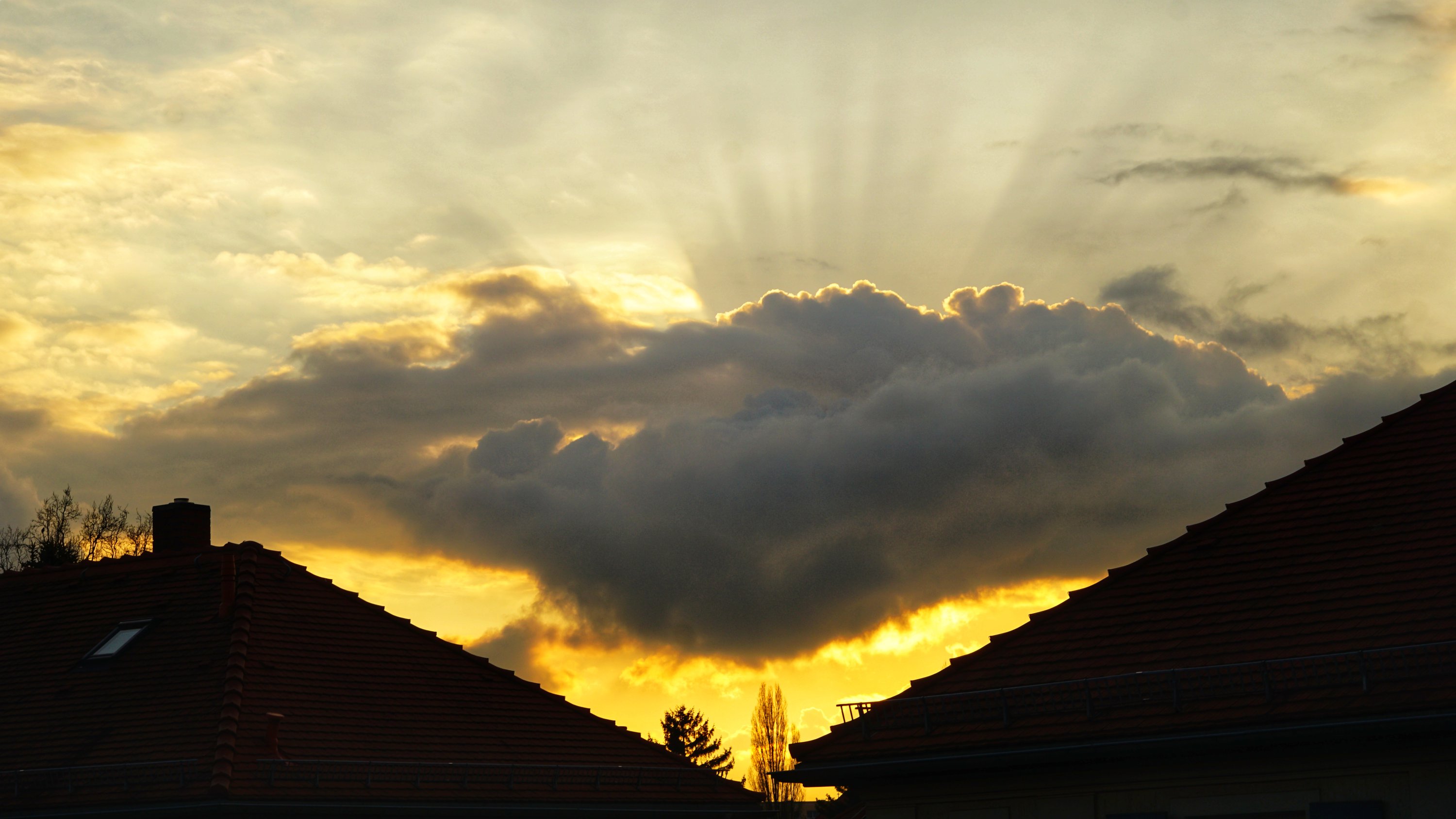 Wolken zwischen Dächern, goldenes Licht am unteren Rand, sichtbare Strahleneffekte oben