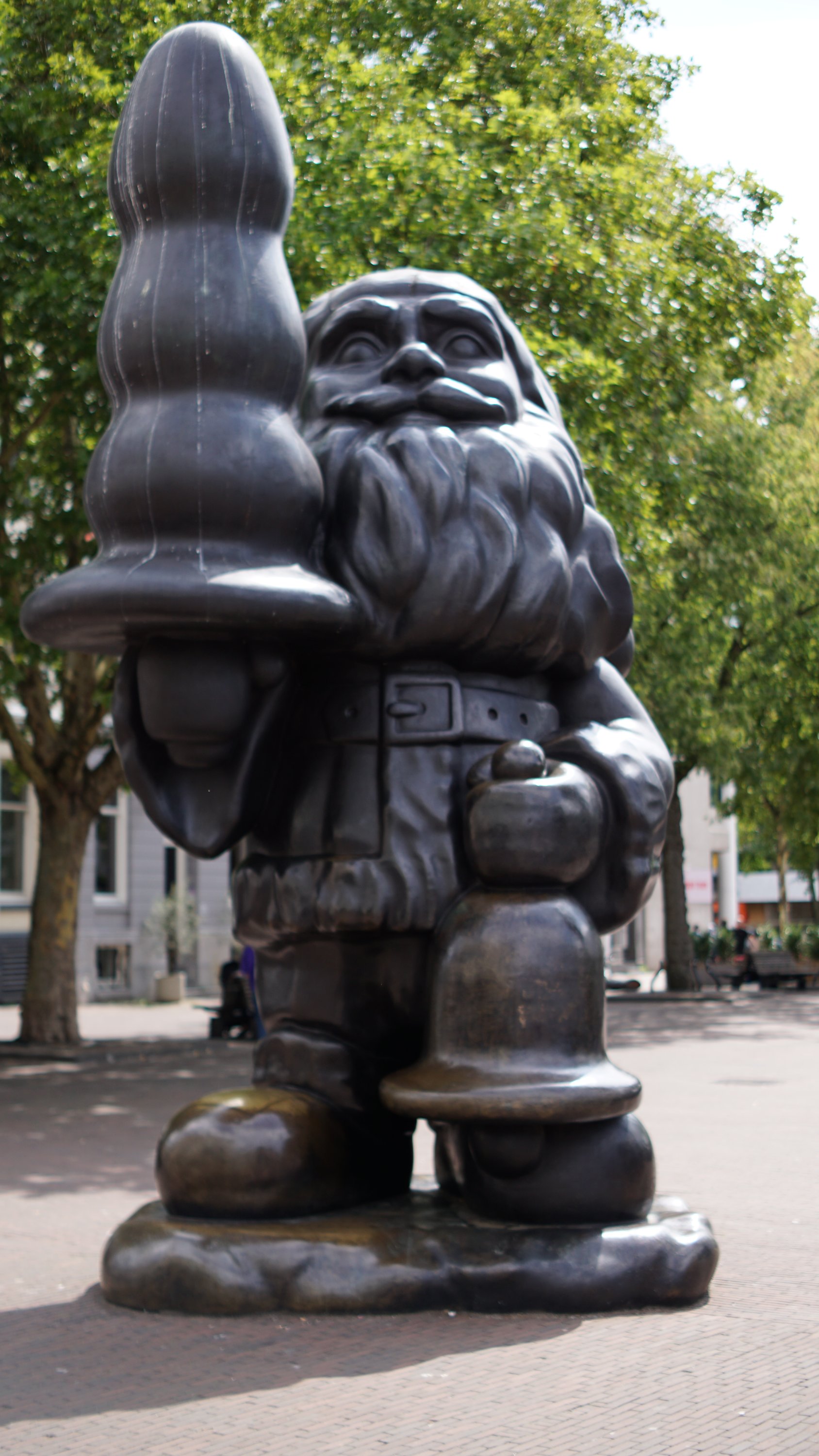 Übergroßer, bärtiger Zwerg (?) hält eine Glocke in der einen Hand und irgendwas knubbeliges, phallisches in eigener Körpergröße in der anderen?

Es könnte einen stilisierten Tannenbaum darstellen - zumindest ist das die einzige, harmlose Erklärung die mir einfällt.

Es ist eine Statue in der Innenstadt von Rotterdam.