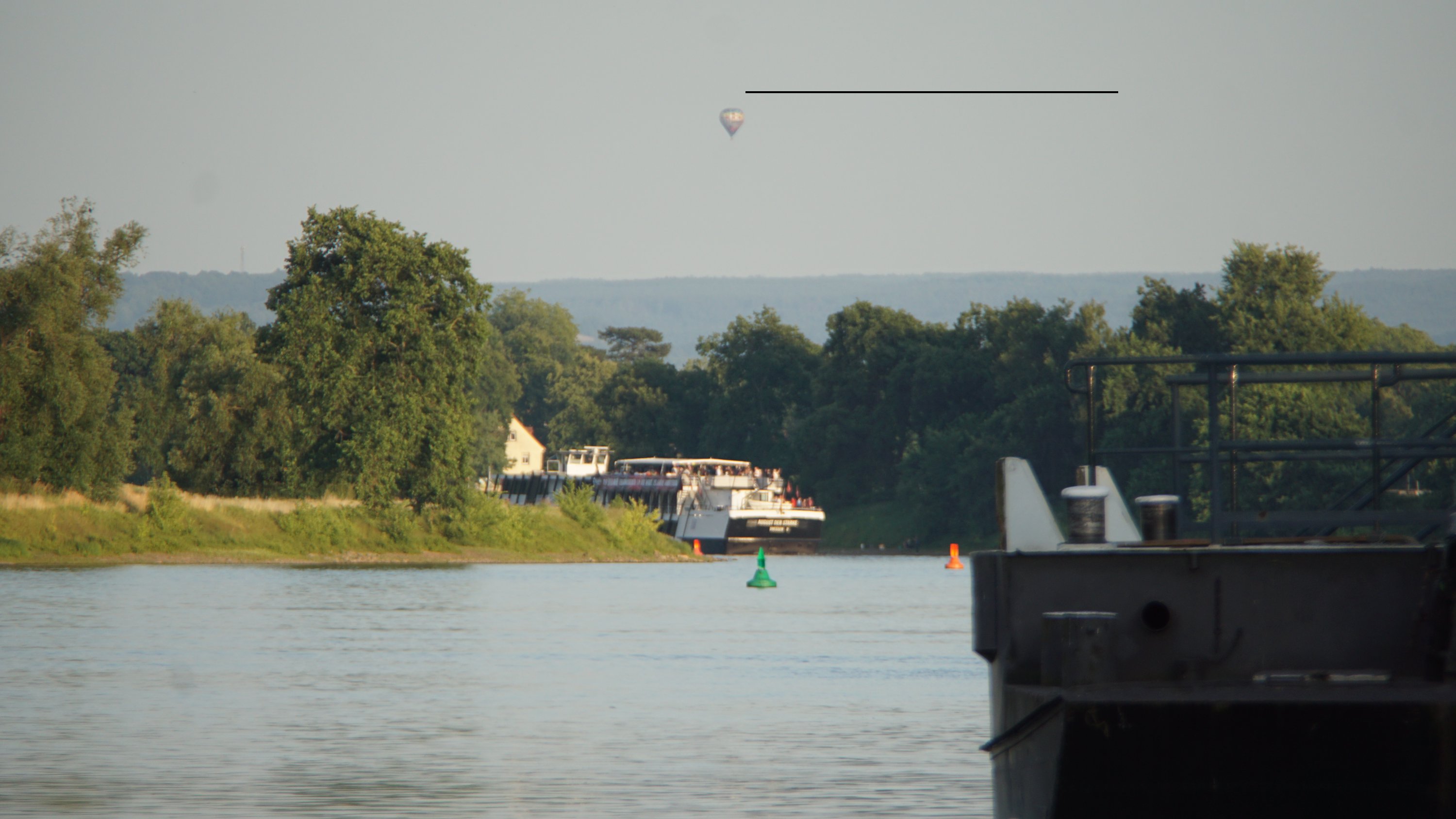 Dampfer verschwindet in der Ferne um eine Kurve im Fluss, Heißluftballon oben drüber, Anleger unscharf im Vordergrund 