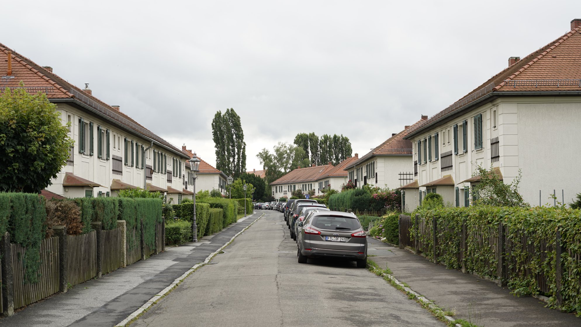 Blick eine schmale Straße entlang. Kleinere Reihenhäuser rechts/links und Autos am Fahrbahnrand.