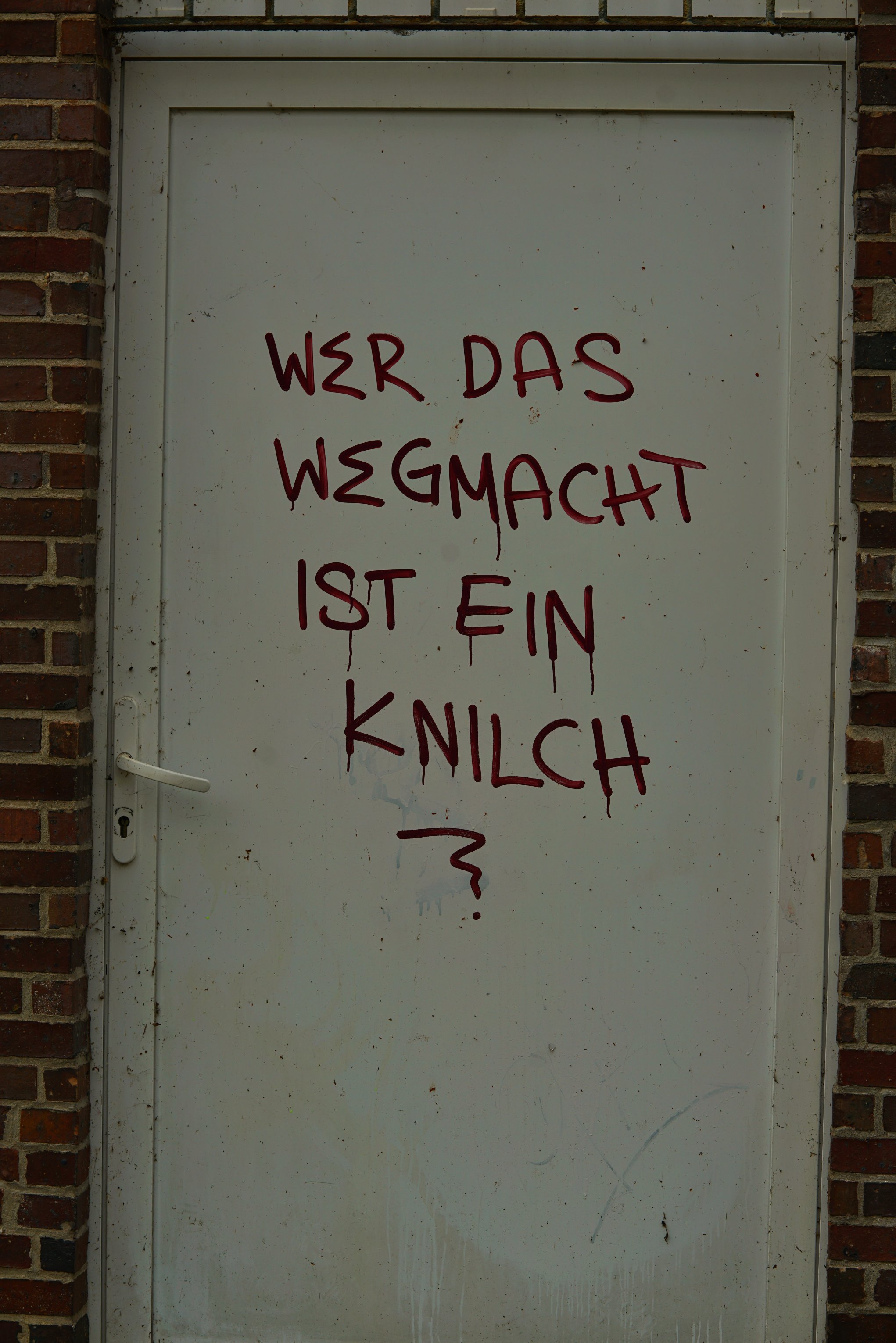 Eine dreckige Blechtür in einer Backsteinwand 

Jemand schmierte "Wer das wegmacht ist ein Knilch?" drauf.