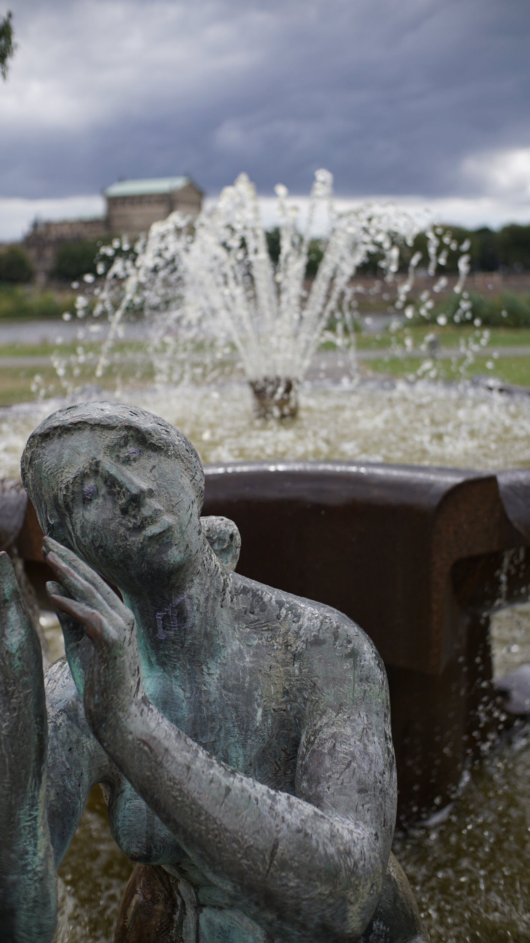 Frauenfigur an einem Brunnen.
Im Hintergrund ist die zugehörige Fontäne zu sehen.
Noch weiter im Hintergrund, erkennt man das Dach der Semperoper, wenn auch sehr unscharf.
Dazwischen liegen Elbe und Grünfläche, diese sind höchstens zu erahnen.