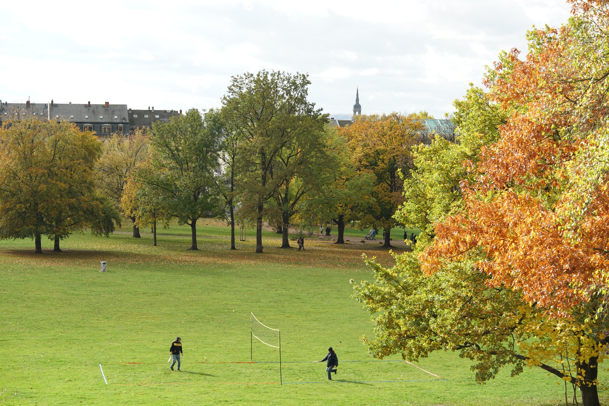 Park mit herbstlichen Bäumen vom Hügel aus aufgenommen. Leute spielen irgendwas Richtung Federball?