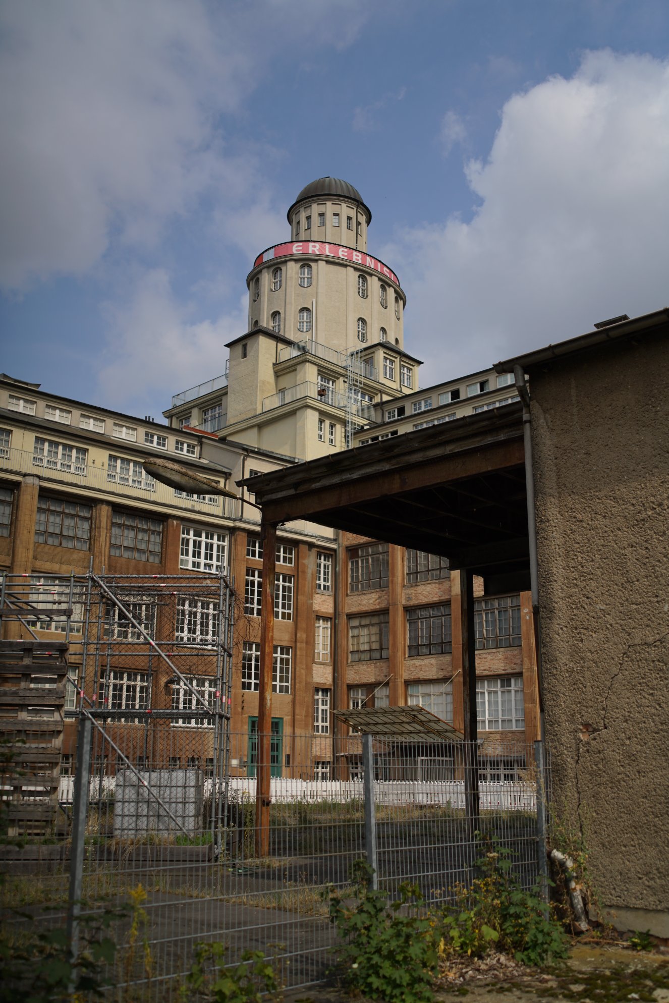 industriebau mit markantem Turm - Sicht von einer (offenen) Ecke des Innenhofes aus, Turm ist auf der gegenüberliegenden Ecke ins Gebäude integriert.

Der Komplex macht einen etwas heruntergekommenen Eindruck, wobei der Teil des Turmes am saubersten aussieht.
Im Hof sind Metallgerüste und Straßensperren gelagert. Eine jahrzehnte alte Straßenlaterne ragt ins Bild.

blauer Himmel und kleine Wölkchen darüber