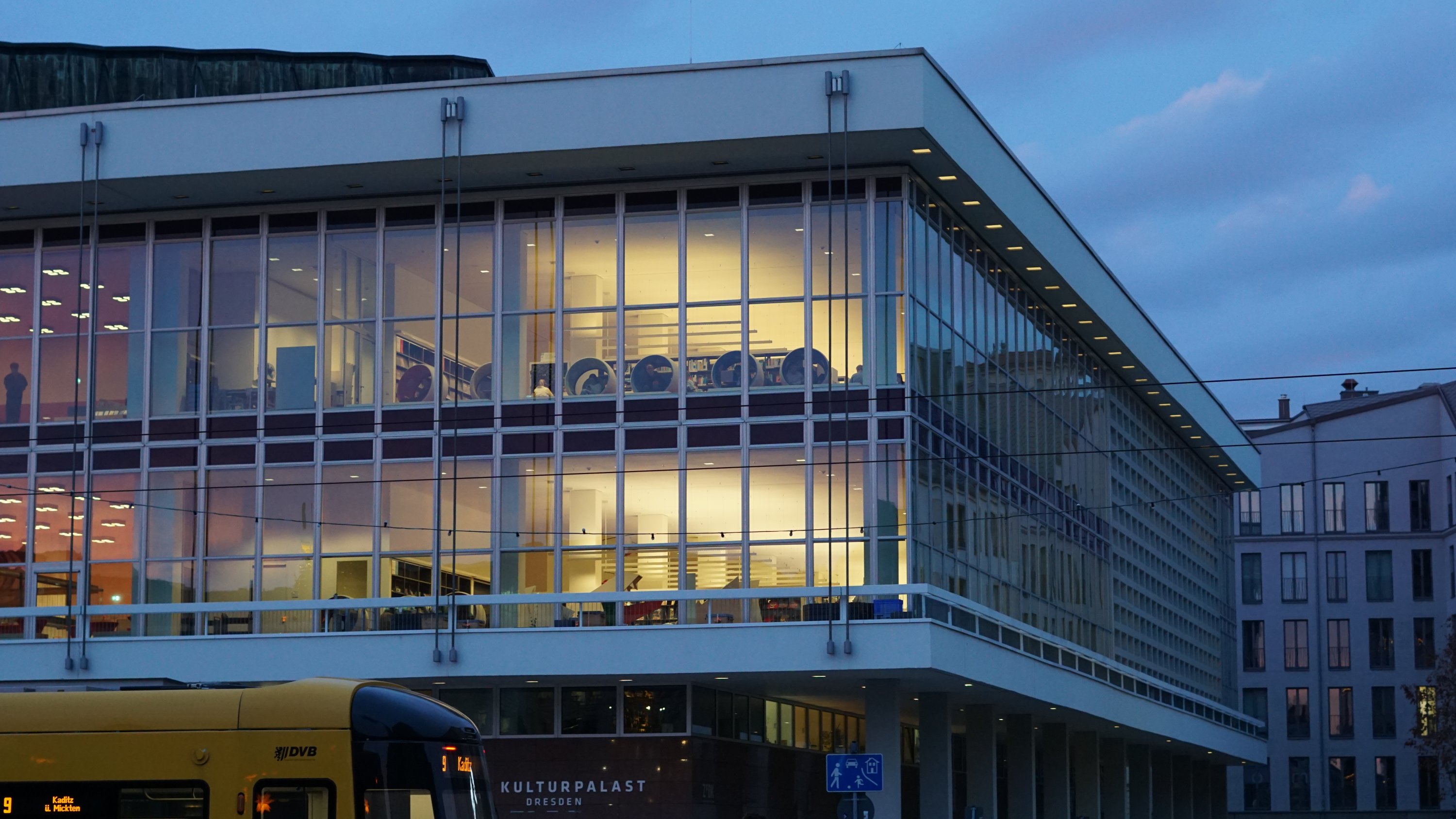 Glasfront des Kulturpalastes mit Stadtbibliothek erleuchtet 
Straßenbahn am vorderen Rand 