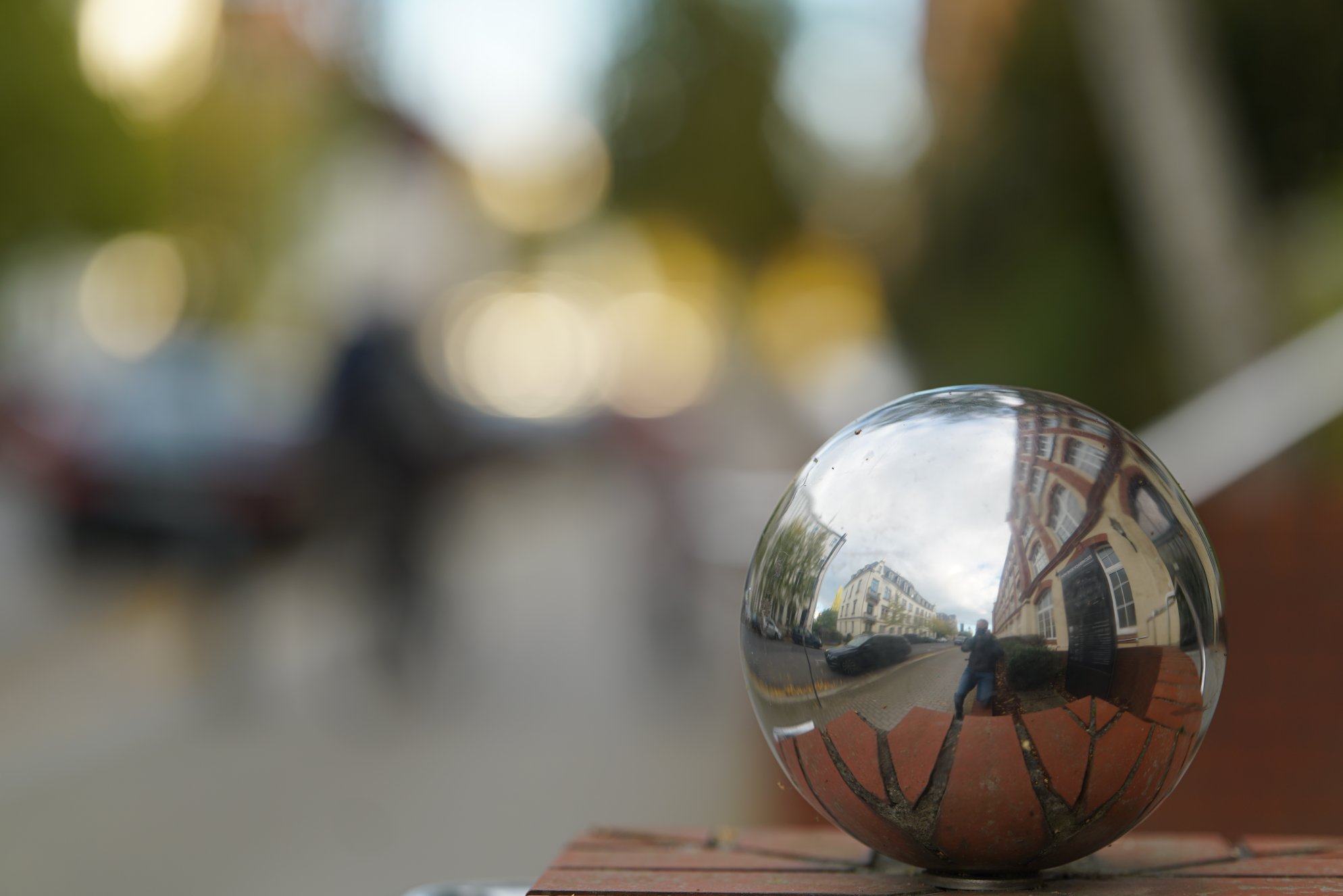 Reflektierende Metallkugel auf Klinker-Mauer, Fotograf spiegelt sich darin, Hintergrund stark unscharf 