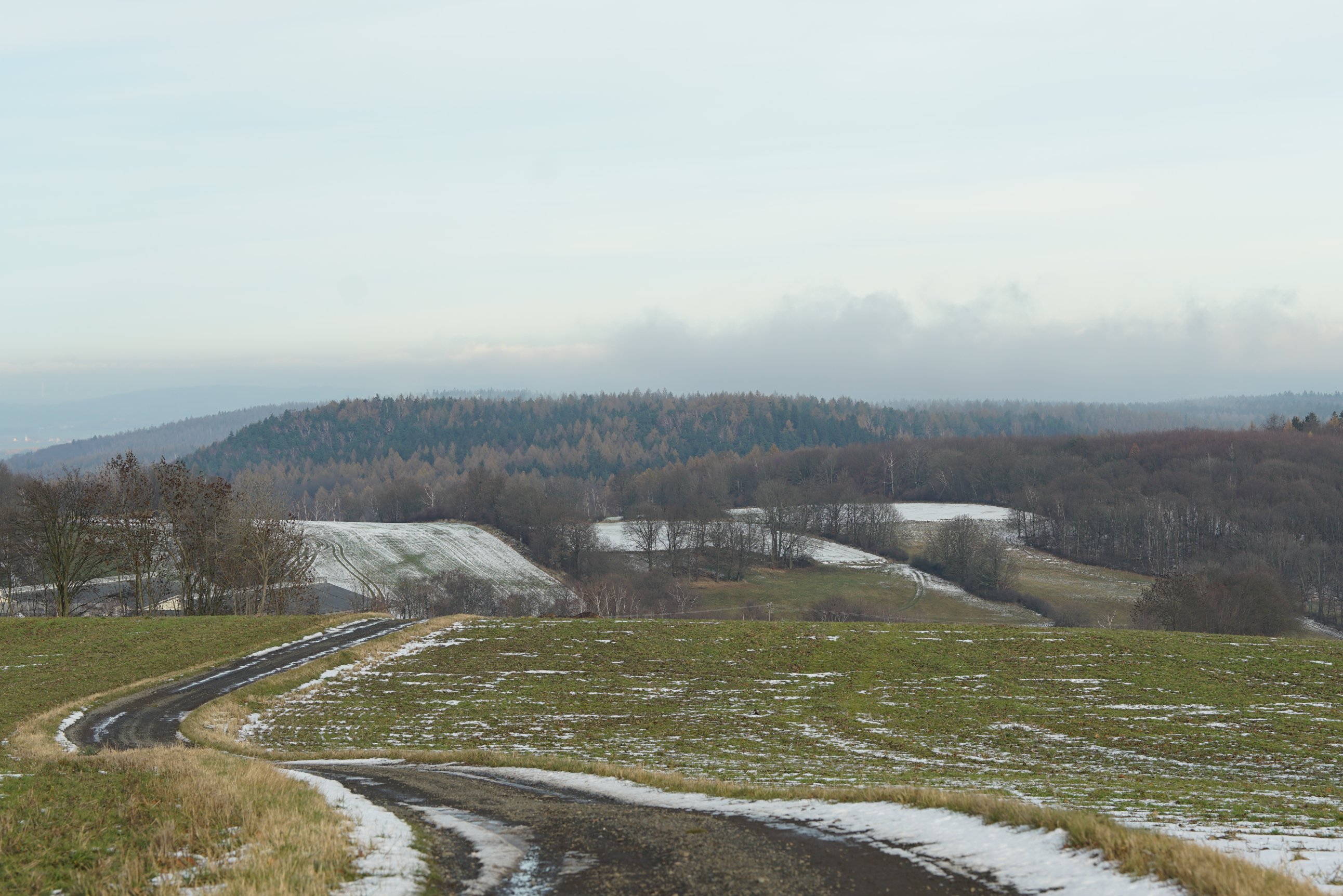 Hügel mit kaum-befestigter Straße, die sich durch die Landschaft windet. Im Hintergrund Wälder und Wolken 