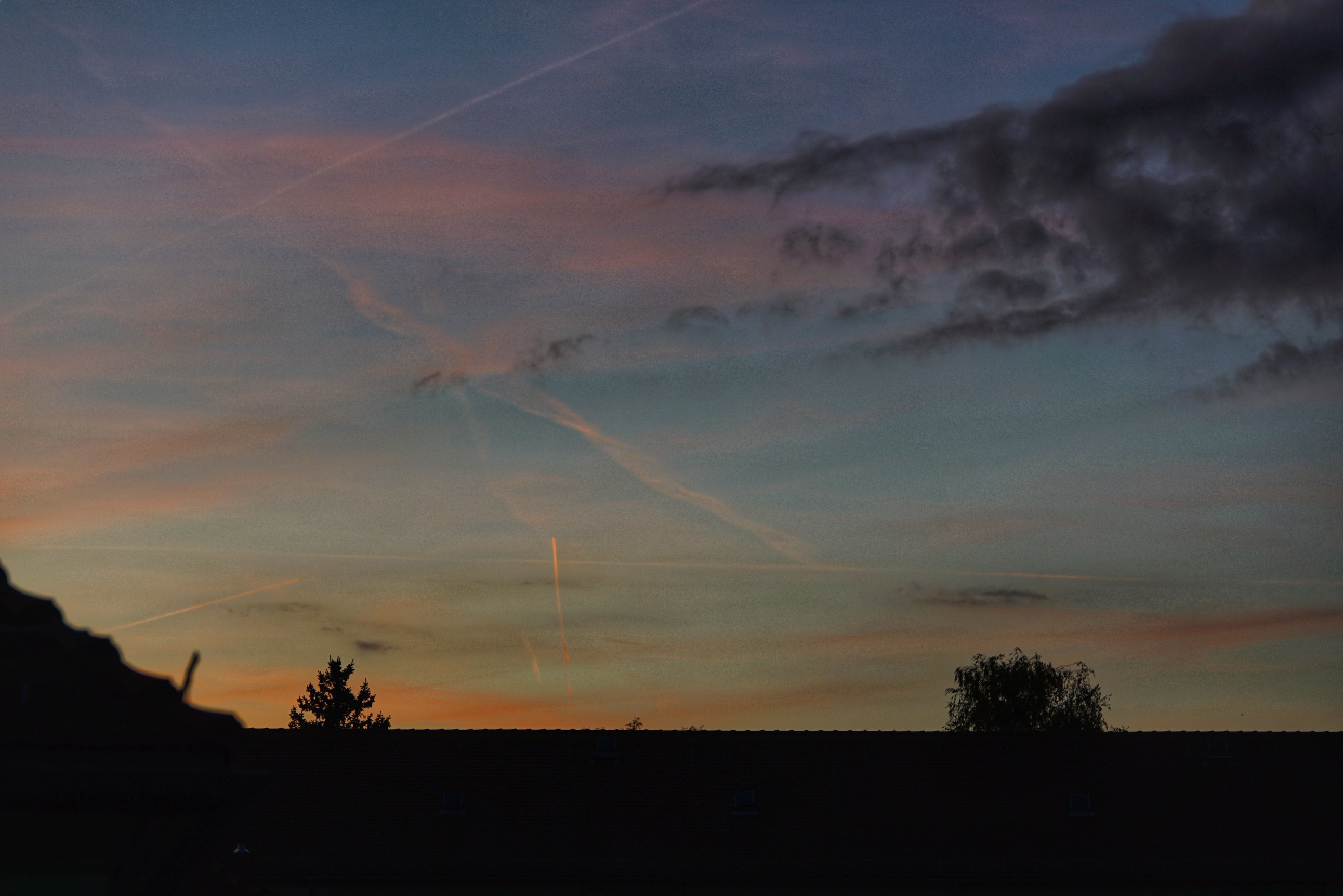 Orange bis rosa Wolken, daneben dunklere. Angeleuchteter Kondensstreifen, Dachspitze und Bäume 